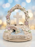 Decorative figurine with cross, flowers, and 'He Is Risen' text against a blurred light background