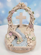 Decorative item with cross, flowers, and 'He Is Risen' text against a blue sky background.