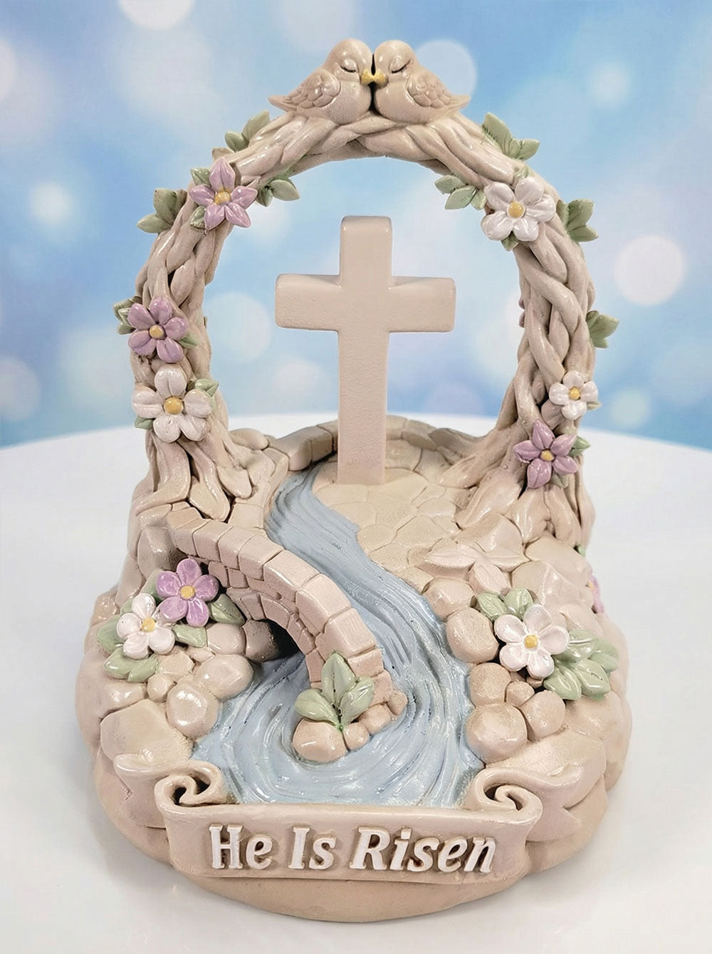 Decorative item with cross, flowers, and 'He Is Risen' text against a blue sky background.