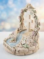 Decorative figurine with cross, flowers, and 'He Is Risen' text against a blurred natural background.