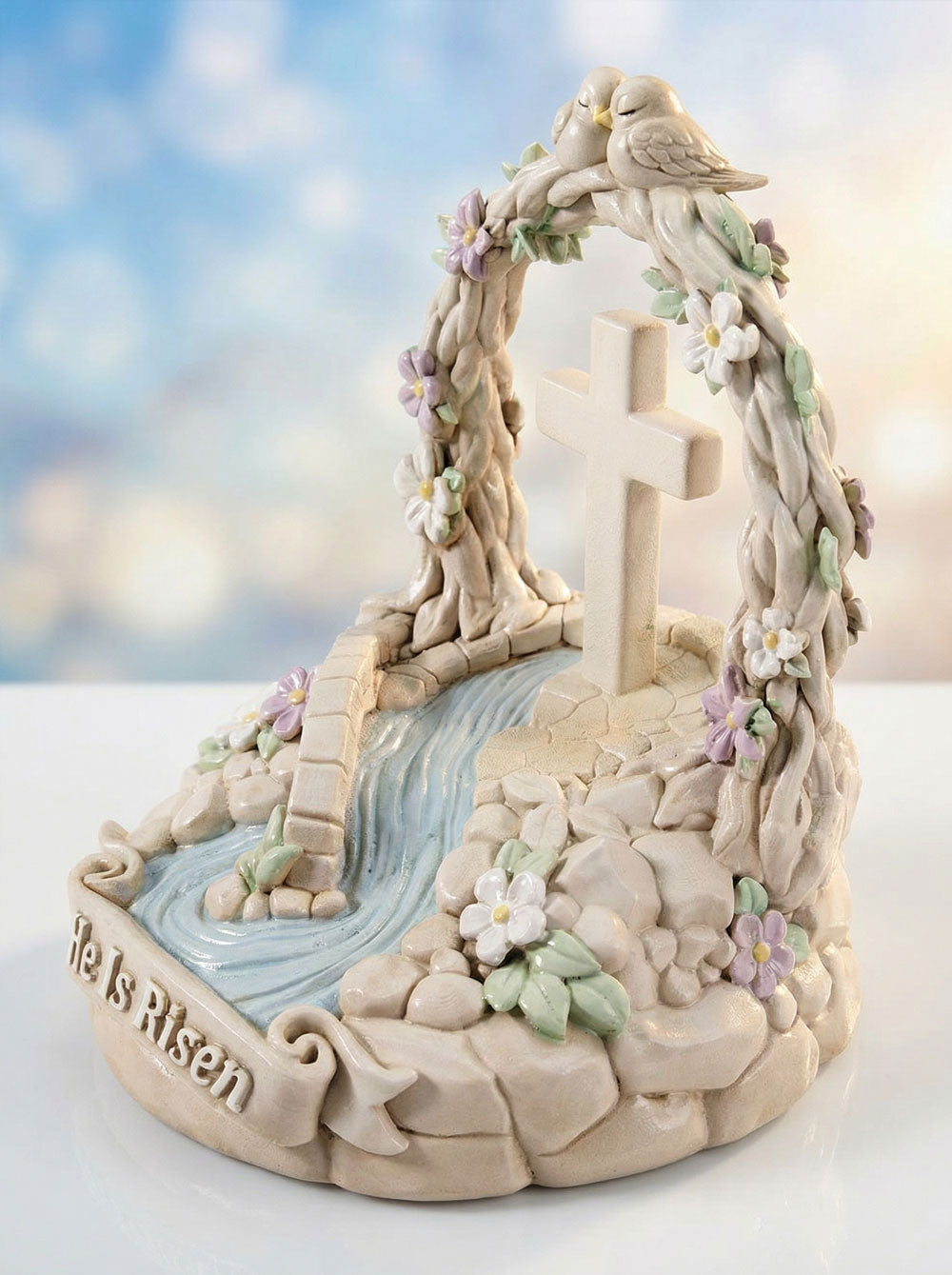Decorative figurine with cross, flowers, and 'He Is Risen' text against a blurred natural background.